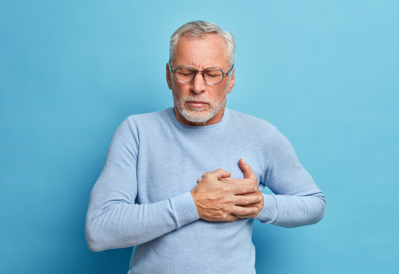 Homem idoso com óculos pressiona a mão no peito, tem ataque cardíaco, sofre de uma dor insuportável, fecha os olhos e usa óculos ópticos poses contra a parede azul, representando as principais doenças cardiovasculares.
