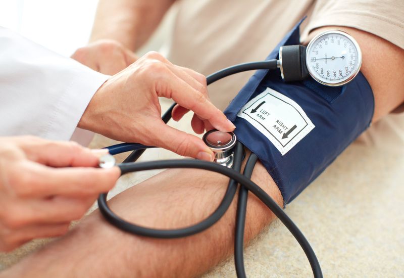 Profissional da área da saúde medindo a pressão arterial de um paciente usando esfigmomanômetro e estetoscópio, representando a hipertensão e a relação com doenças cardiovasculares.