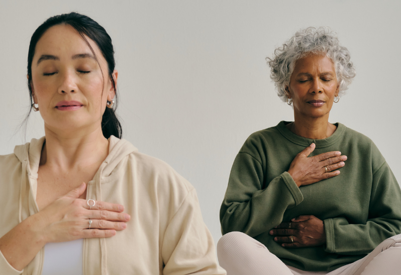 Duas mulheres praticando meditação e mindfulness, focando na respiração consciente, com uma delas tendo cabelos grisalhos e a outra com cabelo escuro, em ambiente tranquilo, representando como a alimentação impacta humor, foco e saúde cardiovascular.